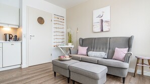 A modern living room with a grey sofa, a matching ottoman, a small side table, and a kitchen area with a kettle and a coffee maker.