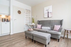 Ein modernes Wohnzimmer mit einem grauen Sofa, passendem Hocker, einem kleinen Beistelltisch und einer Küchenzeile mit Wasserkocher und Kaffeemaschine.