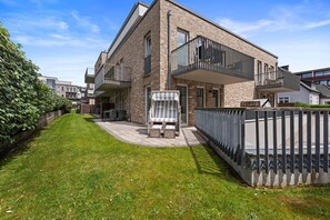 A modern two-story house with a balcony, a small patio with outdoor furniture, and a well-maintained lawn.