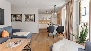 A modern living room with a sofa, coffee table, and chairs, a kitchen in the background, and a large window with curtains. - Beach Suite, App. 2 - Nah am Meer- Sand im Haar und Salz in der Luft (Sylt)