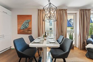 A dining area with a white table set for four, surrounded by dark chairs, a chandelier, and a painting of a bird. - Beach Suite, App. 2 - Nah am Meer- Sand im Haar und Salz in der Luft (Sylt)