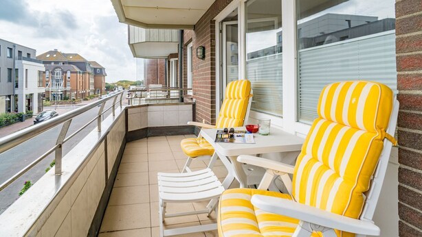 A balcony with yellow-striped chairs and a table, overlooking a street with buildings. - Margaretheneck App. 5 - 2-room vacation apartment close to the beach for max. 2 people (Sylt)