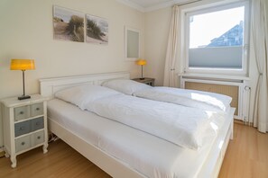 Uma cama de casal com roupa de cama branca, uma mesinha de cabeceira com abajur, um espelho e duas fotos emolduradas na parede.