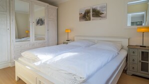 A double bed with white linens, a nightstand with a lamp, a wardrobe, and two wall-mounted picture frames. - Margaretheneck App. 5 - 2-room vacation apartment close to the beach for max. 2 people (Sylt)