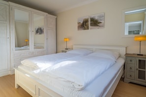 Ein Doppelbett mit weißer Bettwäsche, ein Nachttisch mit Lampe, ein Kleiderschrank und zwei an der Wand befestigte Bilderrahmen.