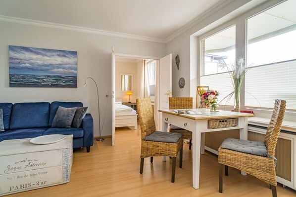 A cozy living room with a blue sofa, a dining table with wicker chairs, and a view of the sea through large windows.