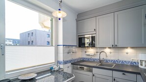 A compact kitchen with a built-in microwave, a sink, and a dishwasher. There is a window with blinds, a decorative blue and white tile backsplash, and a pendant light. - Margaretheneck App. 5 - 2-room vacation apartment close to the beach for max. 2 people (Sylt)