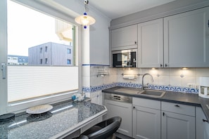 Eine kompakte Küche mit integriertem Mikrowellenherd, Spüle und Geschirrspüler. Ein Fenster mit Jalousien, ein dekorativer, blau-weißer Fliesenspiegel und eine Pendelleuchte sind vorhanden.