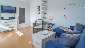 A living room with a blue sofa, a wicker chair, a white cabinet with a motivational quote, a flat-screen TV, and a lamp. - Margaretheneck App. 5 - 2-room vacation apartment close to the beach for max. 2 people (Sylt)
