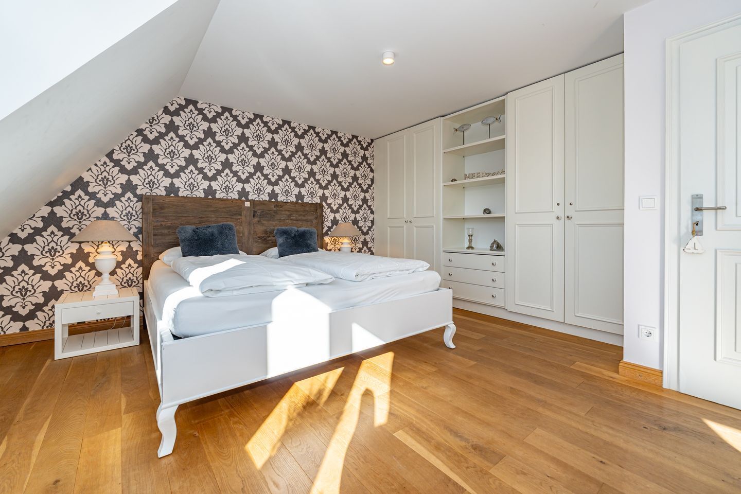 Ein Schlafzimmer mit schrägen Wänden, einem Bett mit weißer Bettwäsche, einem Nachttisch mit Lampe und einem offenen Kleiderschrank.