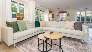Un salon moderne avec un canapé d’angle, deux petites tables rondes et de grandes fenêtres donnant sur un paysage verdoyant.