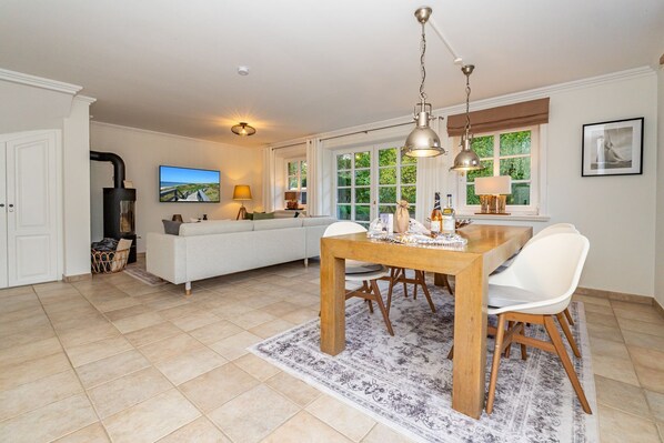 A spacious living room with a sofa, a dining table with chairs, a fireplace, and a large window with a view of greenery. - Lerchennest - vacation home for up to 6 people with garden. - only 500 meters (Sylt)