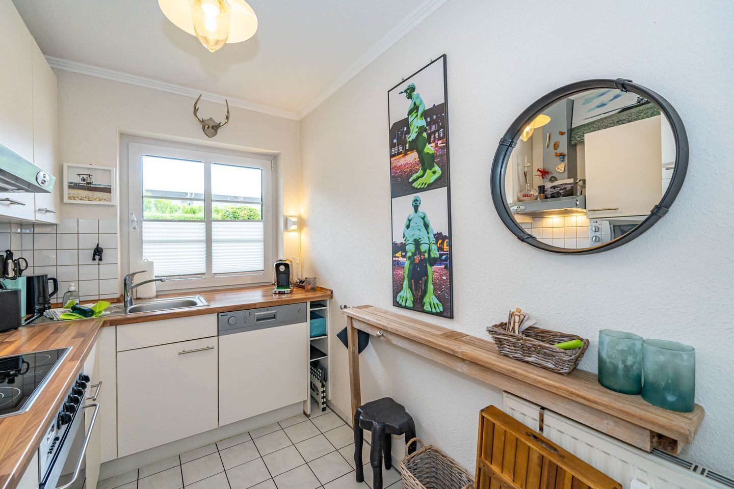A compact kitchen with a dishwasher, stove, and a window with blinds. There are two framed artworks on the wall and a round mirror above the counter.
