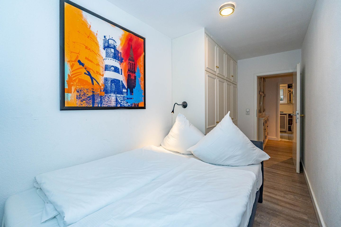 A bedroom with a bed, white pillows, a nightstand, a wall-mounted lamp, and a colorful abstract painting.