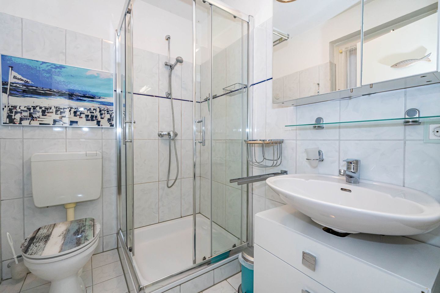 Un baño moderno con ducha envuelta en cristal, inodoro blanco y lavamanos blanco con repisa de vidrio.
