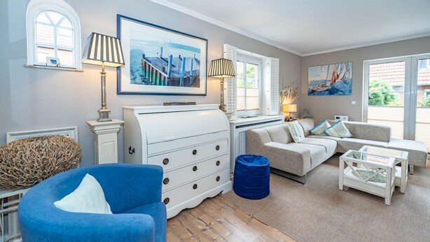 A well-lit living room with a blue armchair, a white dresser, a sofa, and a glass coffee table. There are two windows with shutters, a painting on the wall, and a lamp on a side table. - Constantin - 3-room vacation apartment for up to 4 people with garden and (Sylt)