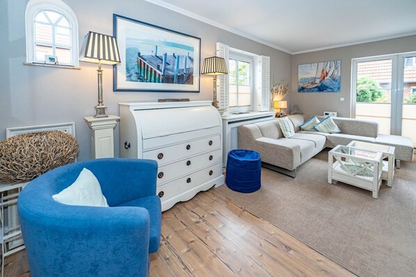 A well-lit living room with a blue armchair, a white dresser, a sofa, and a glass coffee table. There are two windows with shutters, a painting on the wall, and a lamp on a side table.