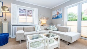 A living room with a sectional sofa, two glass coffee tables, a lamp, a radiator, and a painting of sailboats. - Constantin - 3-room vacation apartment for up to 4 people with garden and (Sylt)