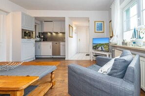 A modern kitchen with white cabinets, a built-in oven, and a dining area with a wooden table and grey sofa. - Strandberg III - 2-room apartment in Westerland - only 400 meters to the beach (Sylt)