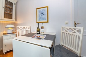 Una habitación pequeña con una mesa blanca, una silla, una lámpara, una botella, un cuadro enmarco y un aparador con cristalería.
