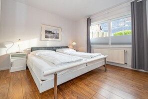 Un lit double avec une tête de lit, deux tables de chevet avec des lampes, un tableau encadré au mur, une fenêtre avec des rideaux et un radiateur.