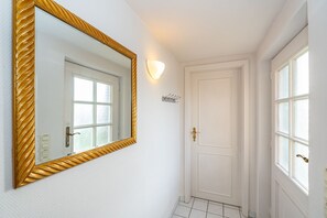 Un couloir avec une porte blanche, un miroir encadré d’or et un luminaire fixé au mur.