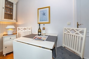 Et lille rum med et hvidt bord, en stol, en lampe, en flaske, et indrammet billede og et skab med glas.