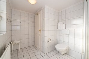 Un baño con paredes de azulejos blancos, un inodoro, una ducha y una mesita con una toalla.