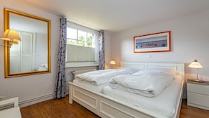 Een slaapkamer met een tweepersoonsbed, een grote spiegel, een raam met gordijnen, een nachtkastje met een lamp en een lijst met een afbeelding aan de muur.