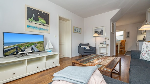 Een woonkamer met een tv waarop een strand te zien is, een houten salontafel, een grijze bank en een grijze fauteuil.