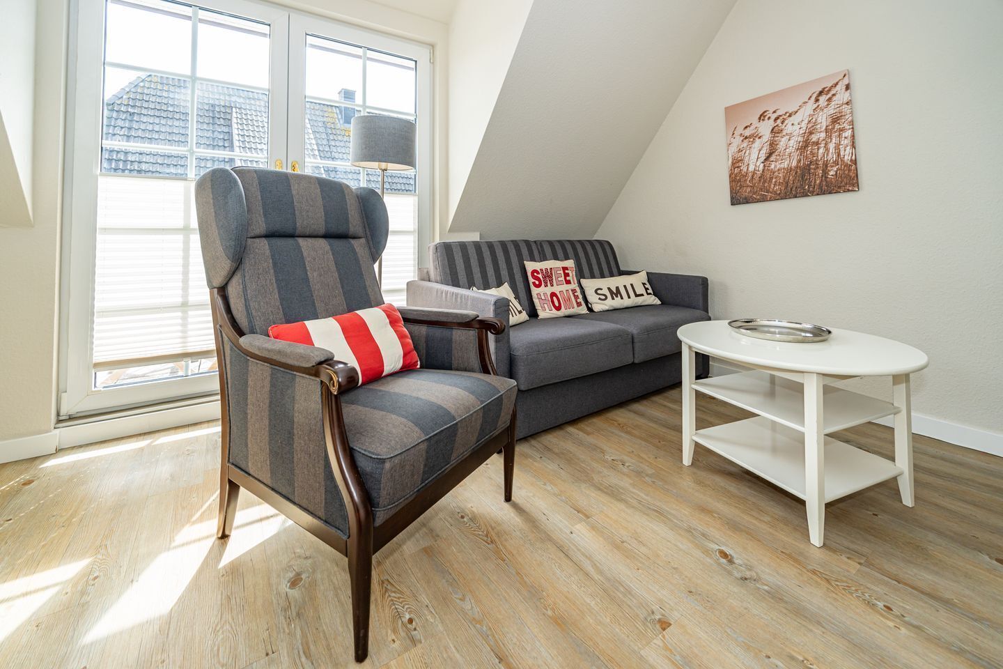 Un salon moderne avec un plafond haut, un canapé gris agrémenté de coussins à rayures, un fauteuil en bois avec un coussin rouge et blanc, et une table basse blanche sur laquelle repose un bol en métal.