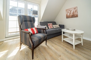 Ein modernes Wohnzimmer mit hoher Decke, ein graues Sofa mit gestreiften Kissen, ein Holzschaukelstuhl mit rotem und weißem Kissen und ein weißer Couchtisch mit einer Metallschale.