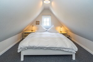 Quarto com cama branca, duas mesas de cabeceira com luminárias, uma janela e uma pequena placa na parede.