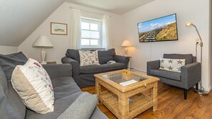 Una sala de estar acogedora con un sofá, un sillón y una mesita de centro. Se aprecia una ventana con cortinas, una lámpara y un cuadro en la pared.