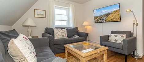 Un salon chaleureux avec un canapé, un fauteuil et une table basse. Il y a une fenêtre avec des rideaux, une lampe et un tableau au mur.