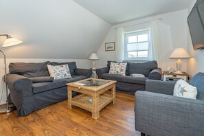 Ein gemütliches Wohnzimmer mit zwei grauen Sofas, einem Korbtisch und einem kleinen Beistelltisch mit Lampe.