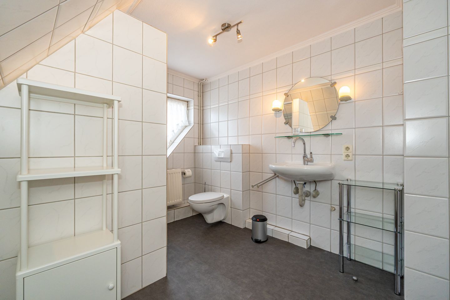 Ein modernes Badezimmer mit weißen Fliesen, einer Glasablage, einer weißen Toilette und einem Waschbecken mit Spiegel darüber.