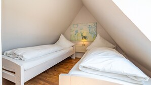Un pequeño dormitorio abuhardillado con forma de triángulo, con dos camas blancas, una mesita de noche con lámpara y un cuadro colorido en la pared.