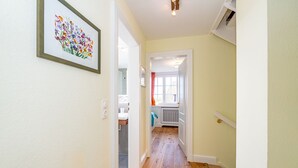 A hallway with wooden flooring, a framed floral artwork on the wall, and a view into a room with a window and a radiator. - Wiip Hüs - Unter Reet Gebautes Hausteil in Keitum, mit Garten für bis zu 4 Personen (Sylt-Ost)