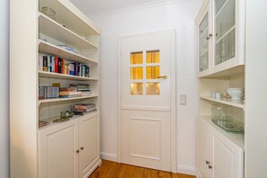 Un couloir étroit avec des meubles blancs, une porte dotée de panneaux de verre et une étagère où sont posés des livres et des objets décoratifs.