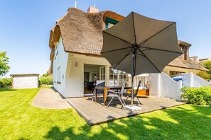 A house with a thatched roof, a wooden deck, and a patio area with a table and chairs. - Wiip Hüs - Unter Reet Gebautes Hausteil in Keitum, mit Garten für bis zu 4 Personen (Sylt-Ost)