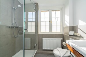 A modern bathroom with a glass shower enclosure, a toilet with a wooden vanity, and a radiator under a window with blinds. - Wiip Hüs - Unter Reet Gebautes Hausteil in Keitum, mit Garten für bis zu 4 Personen (Sylt-Ost)