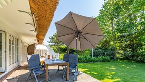 An outdoor patio with a table and chairs, a large umbrella, and a thatched roof structure. - Wiip Hüs - Unter Reet Gebautes Hausteil in Keitum, mit Garten für bis zu 4 Personen (Sylt-Ost)