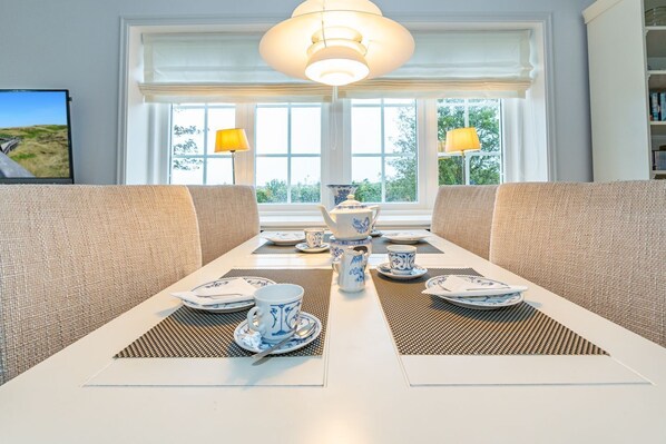 A dining area with a white table set for four, two lamps, and a view of greenery outside.