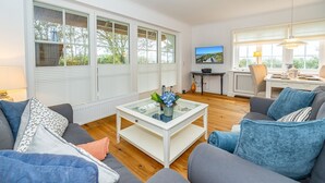 A modern living room with a glass coffee table, grey sofas, and a television on a stand. - Wiip Hüs - Unter Reet Gebautes Hausteil in Keitum, mit Garten für bis zu 4 Personen (Sylt-Ost)