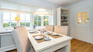 A dining area with a white table set for four, surrounded by chairs, with a window and bookshelf in the background. - Wiip Hüs - Unter Reet Gebautes Hausteil in Keitum, mit Garten für bis zu 4 Personen (Sylt-Ost)