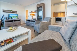 Un salon moderne avec une table basse blanche, un canapé gris à revêtement en tissu et un petit pouf marron. Un téléviseur à écran plat est fixé au mur, et on aperçoit un coin cuisine à l’arrière-plan.