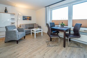 Een moderne woonkamer met een grijze bank, een witte salontafel en een eettafel met vier stoelen. Op de muur hangt een schilderij, er staat een vloerlamp en er is een raam met jaloezieën.