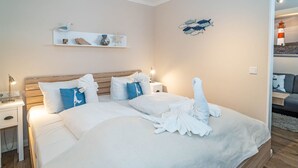 A neatly made bed with white linens and a towel arranged on the bed. There is a nightstand with a lamp and a small table beside the bed. A shelf with decorative items is mounted on the wall above the bed. - Strandmuschel App. 309 - Kleine, Geschmackvoll Eingerichtete Wohnung in Westerland, Direkt am (Sylt)