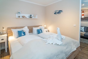 A neatly made bed with white linens and a towel arranged on the bed. There is a nightstand with a lamp and a small table beside the bed. A shelf with decorative items is mounted on the wall above the bed.